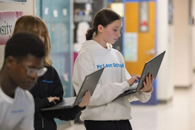 Three students stand in a school hallway holding and using laptops. The student in front is wearing a white McLean School hoodie.