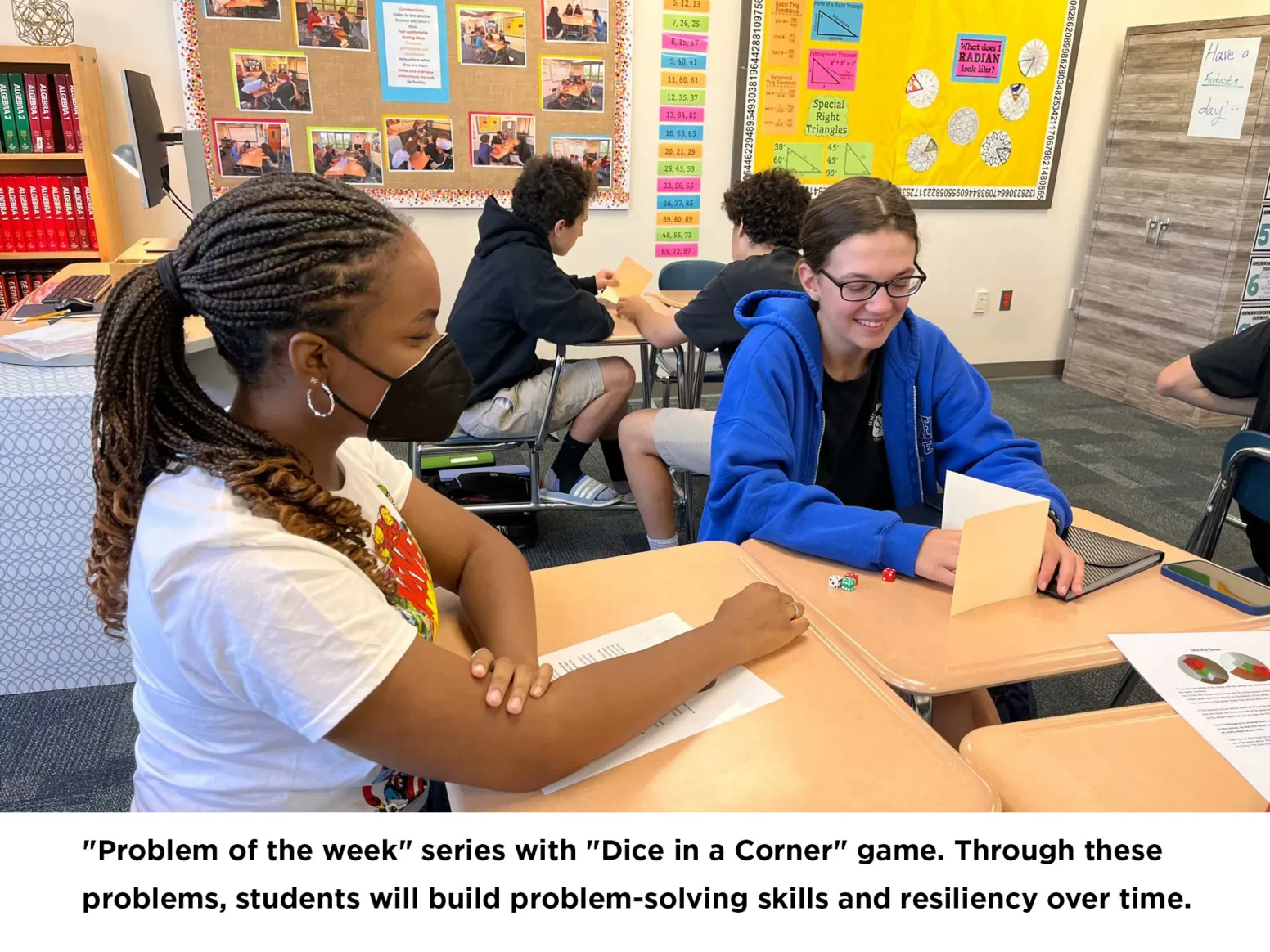 Two students sit at adjoining desks, playing a dice game and discussing a worksheet. Classroom materials and other students are visible in the background.
