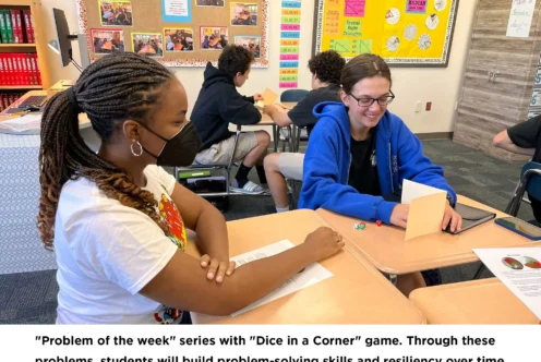 Two students sit at adjoining desks, playing a dice game and discussing a worksheet. Classroom materials and other students are visible in the background.