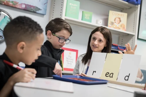 A teacher helps two young boys with a reading activity, using letter cards to spell "read" and a tray of red sand for tactile learning.