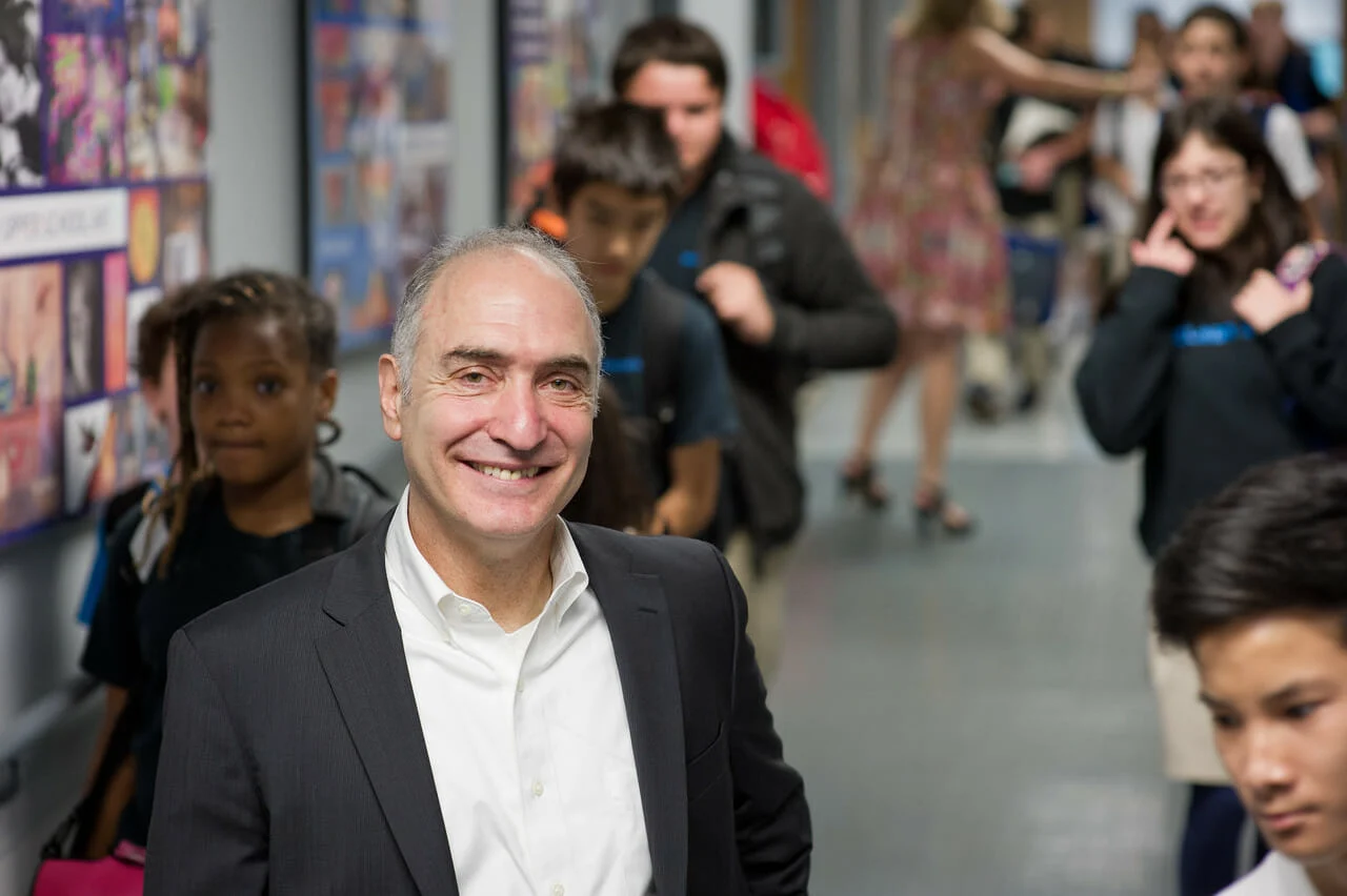 A man in a suit smiles in a school hallway with children and adults in the background.