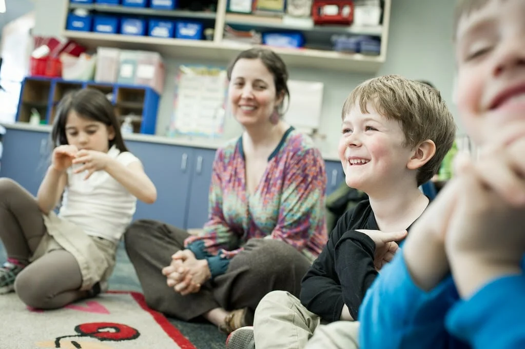 A teacher and three young children sit in a circle on the classroom floor, smiling and interacting during an activity.