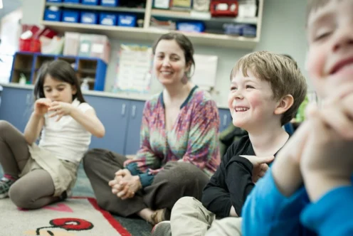 A teacher and three young children sit in a circle on the classroom floor, smiling and interacting during an activity.