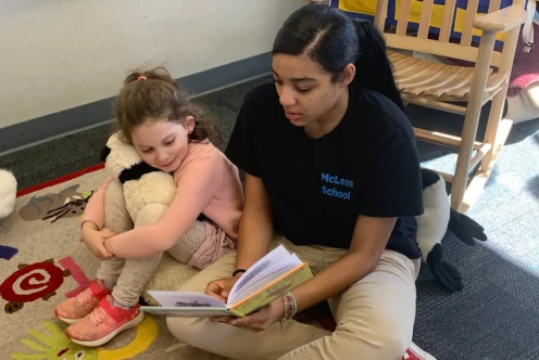 An older student reads a book to a younger student who is hugging a stuffed animal while sitting on a classroom rug.