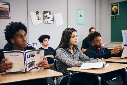 Students sitting at desks in a classroom read textbooks and take notes while facing the front of the room.