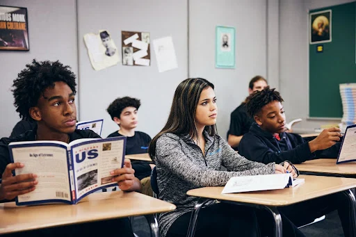 Students sitting at desks in a classroom read textbooks and take notes while facing the front of the room.