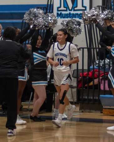 A basketball player in a Mustangs uniform runs onto the court, greeted by cheerleaders and teammates, while spectators watch from the bleachers.