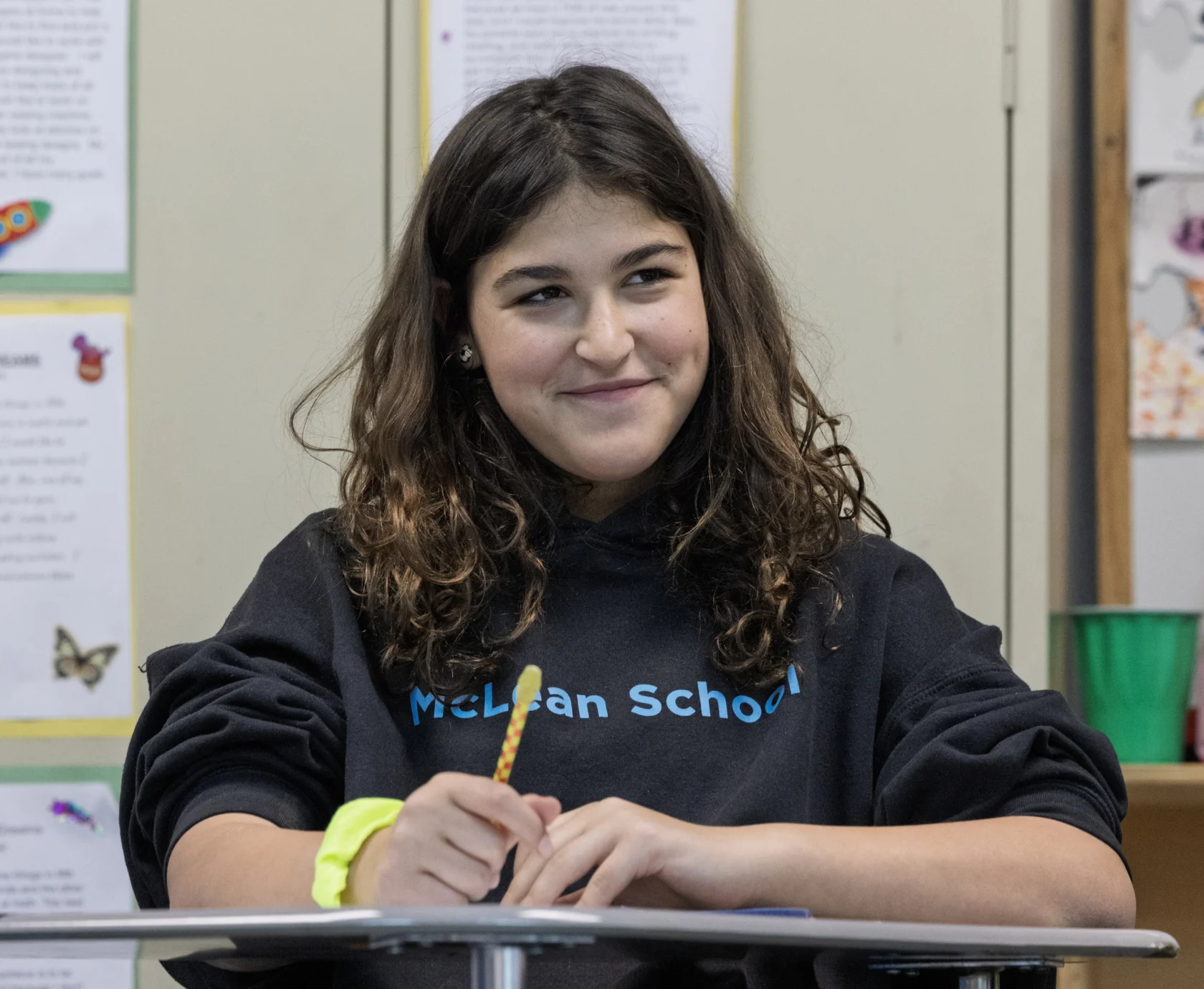 A student wearing a "McLean School" sweatshirt sits at a desk, holding a pencil and smiling, with papers and classroom materials in the background.
