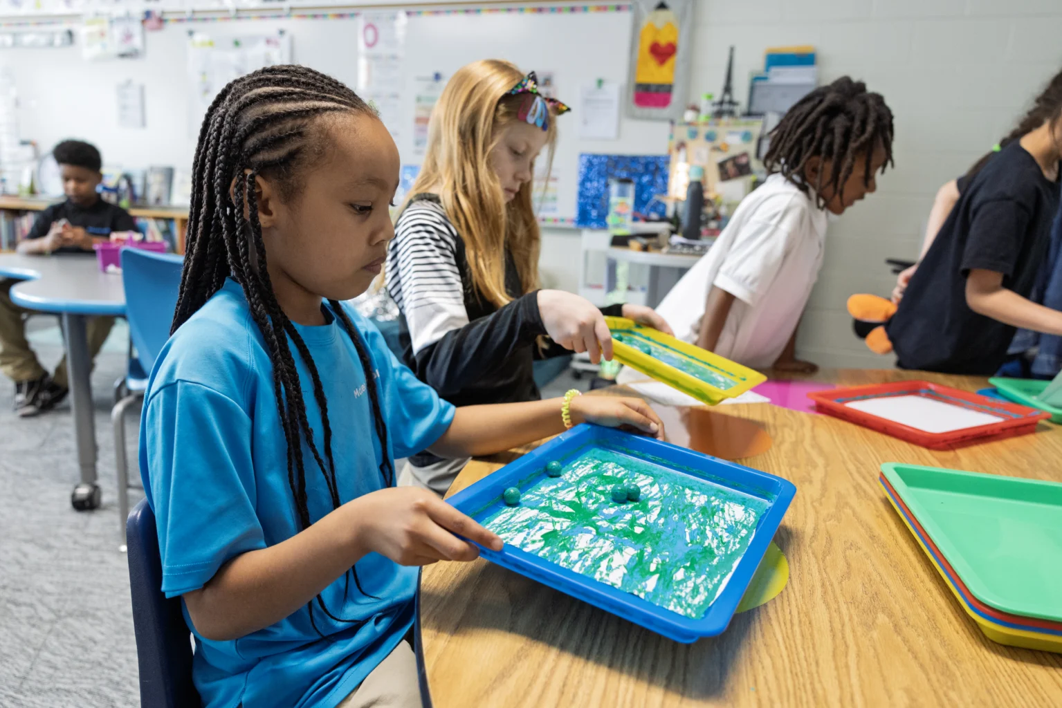 Three children sit at a classroom table, tilting trays filled with green paint, likely engaged in an art or science activity. Other students work in the background.