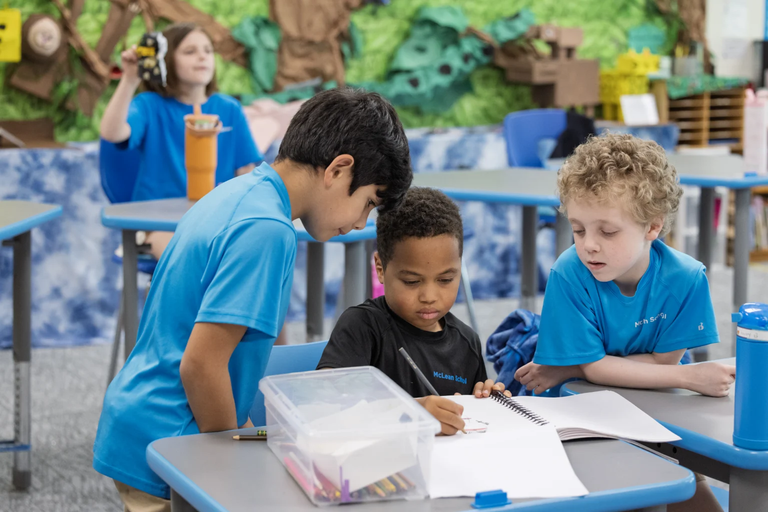 Three boys work together at a desk, looking at a notebook, while another child sits in the background holding up a toy in a colorful classroom.