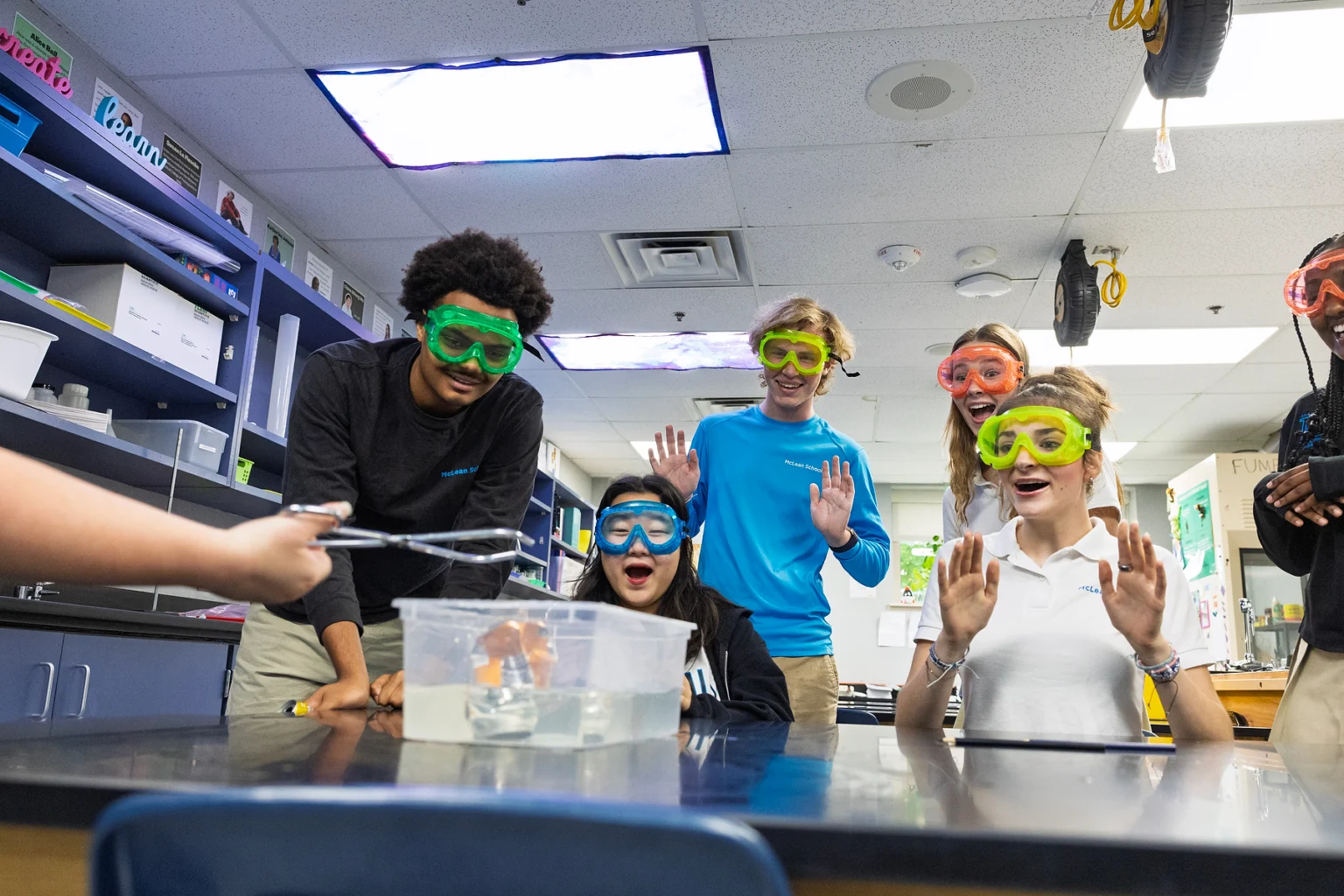 Five students wearing safety goggles react with excitement as one student uses tongs to handle an object in a water-filled container during a science experiment in a classroom.