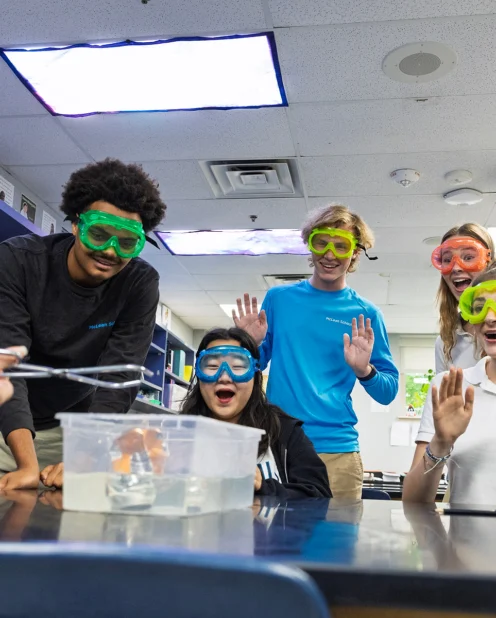 Five students wearing safety goggles react with excitement as one student uses tongs to handle an object in a water-filled container during a science experiment in a classroom.
