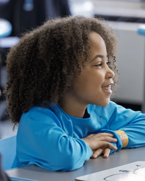 A child with curly hair and a blue shirt sits at a desk, smiling and looking forward in a classroom setting. Another student is partially visible in the foreground.