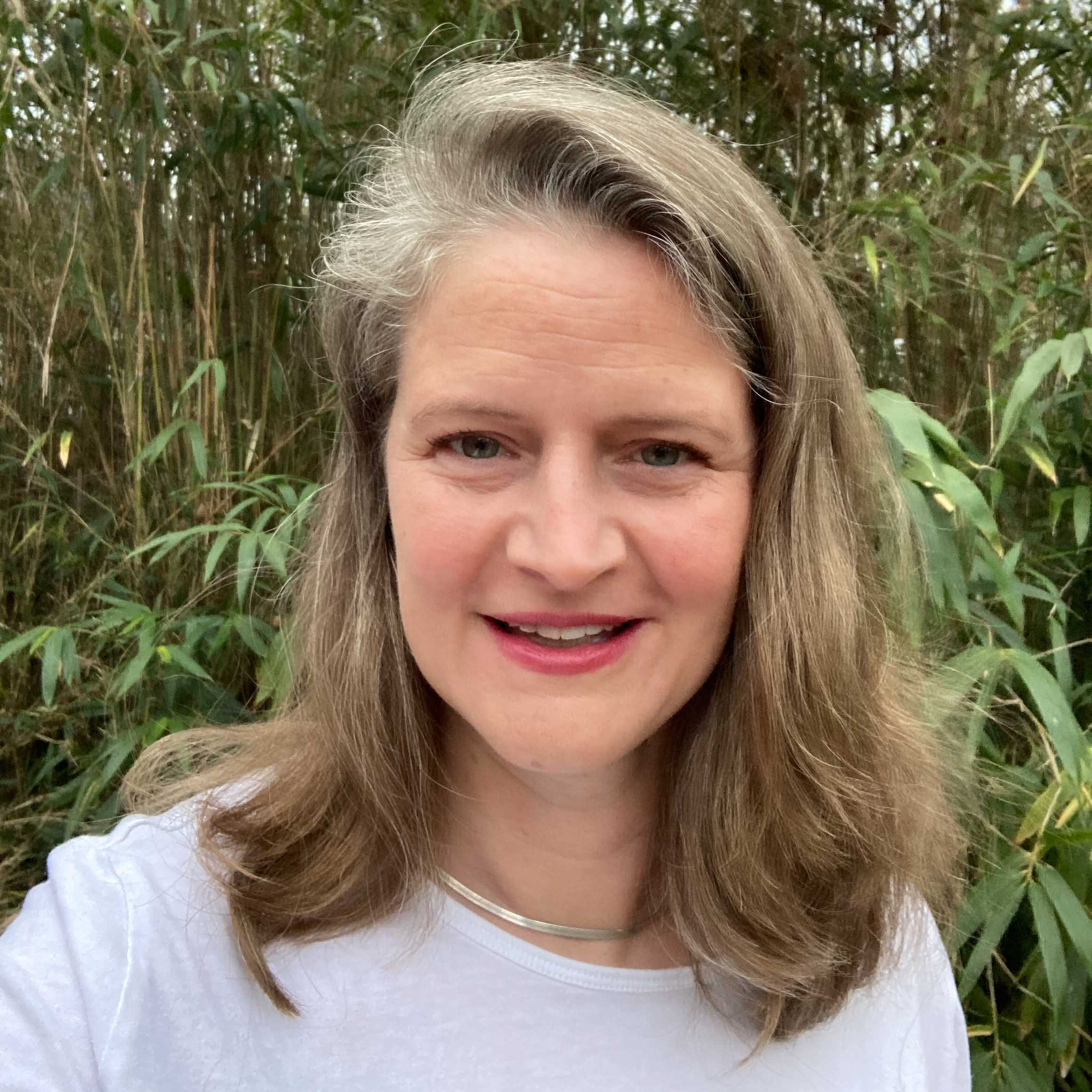 A woman with shoulder-length light brown hair, wearing a white top and silver necklace, stands outdoors in front of green bamboo plants, smiling at the camera.