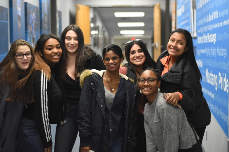 Seven young women stand closely together, smiling at the camera in a brightly lit hallway with blue walls covered in text.