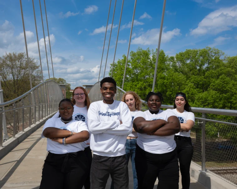Six young people stand together on a bridge, arms crossed, posing for a group photo on a sunny day. They wear white shirts and jeans, with trees and blue sky in the background.