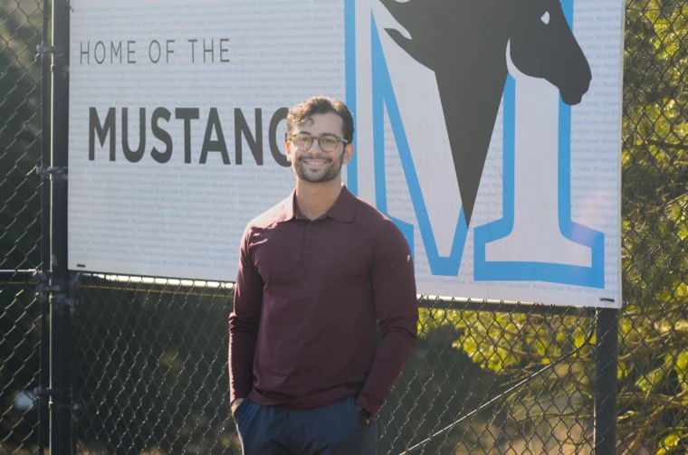 A man wearing glasses and a maroon long-sleeve shirt stands in front of a sign that reads "Home of the Mustangs" with a mustang logo.