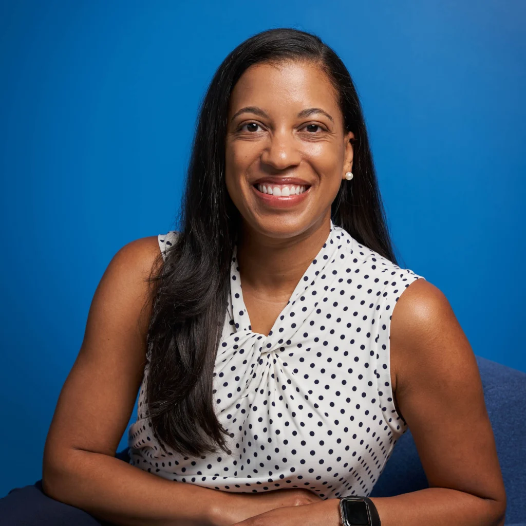 Woman with long dark hair wearing a white sleeveless blouse with black polka dots, sitting and smiling in front of a blue background.