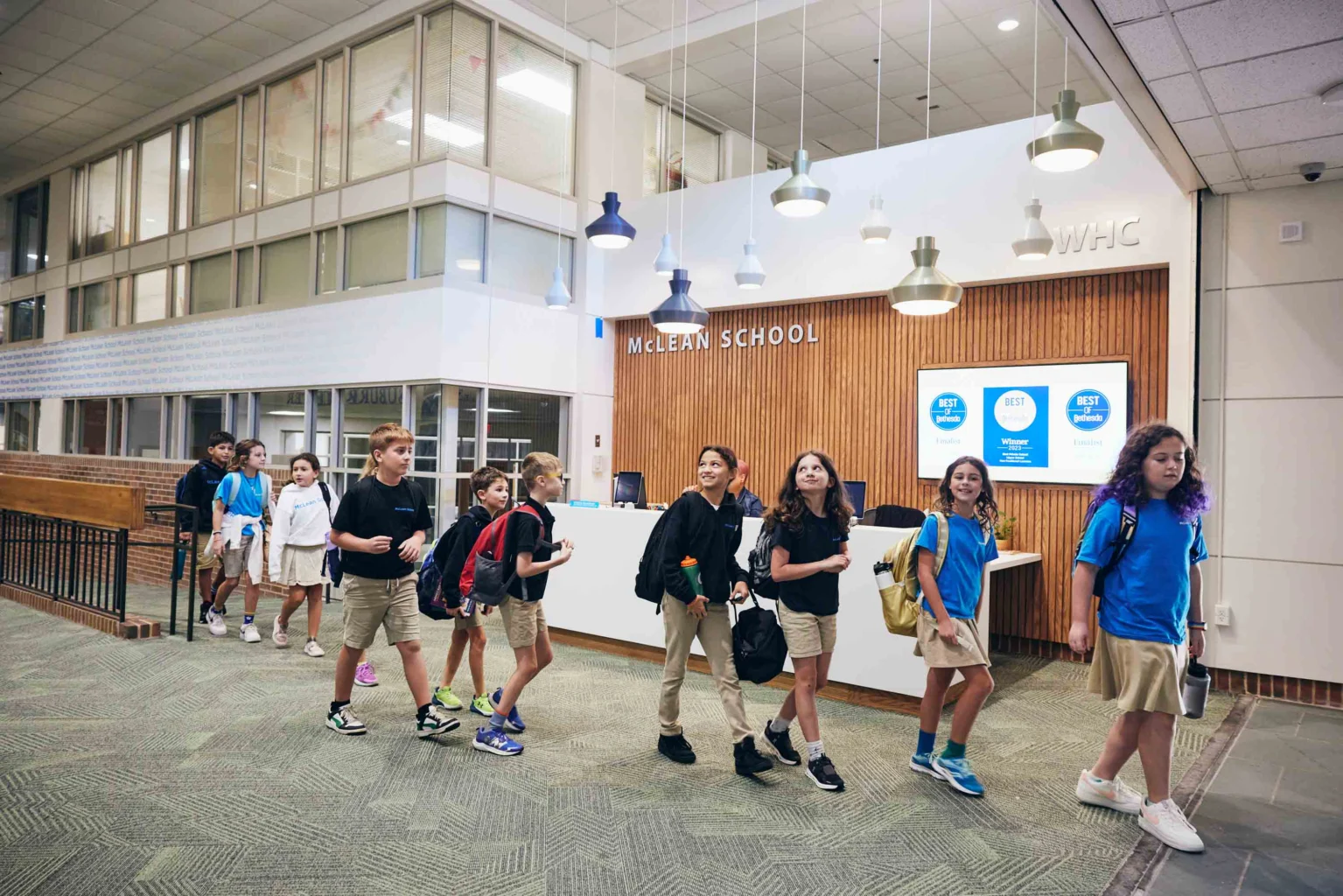 A group of students in uniforms walk in line through a school lobby with a reception desk and "McLEAN SCHOOL" sign on the wall.