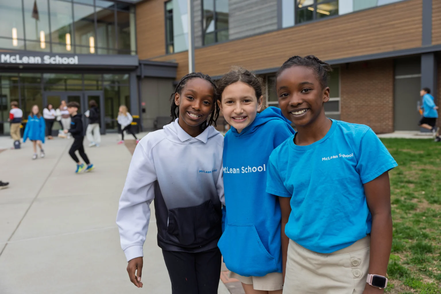 Three students stand smiling in front of McLean School, with other students visible in the background outside the school building.