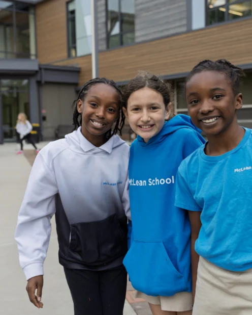 Three students stand and smile outside the McLean School building, with other children visible in the background on the sidewalk.