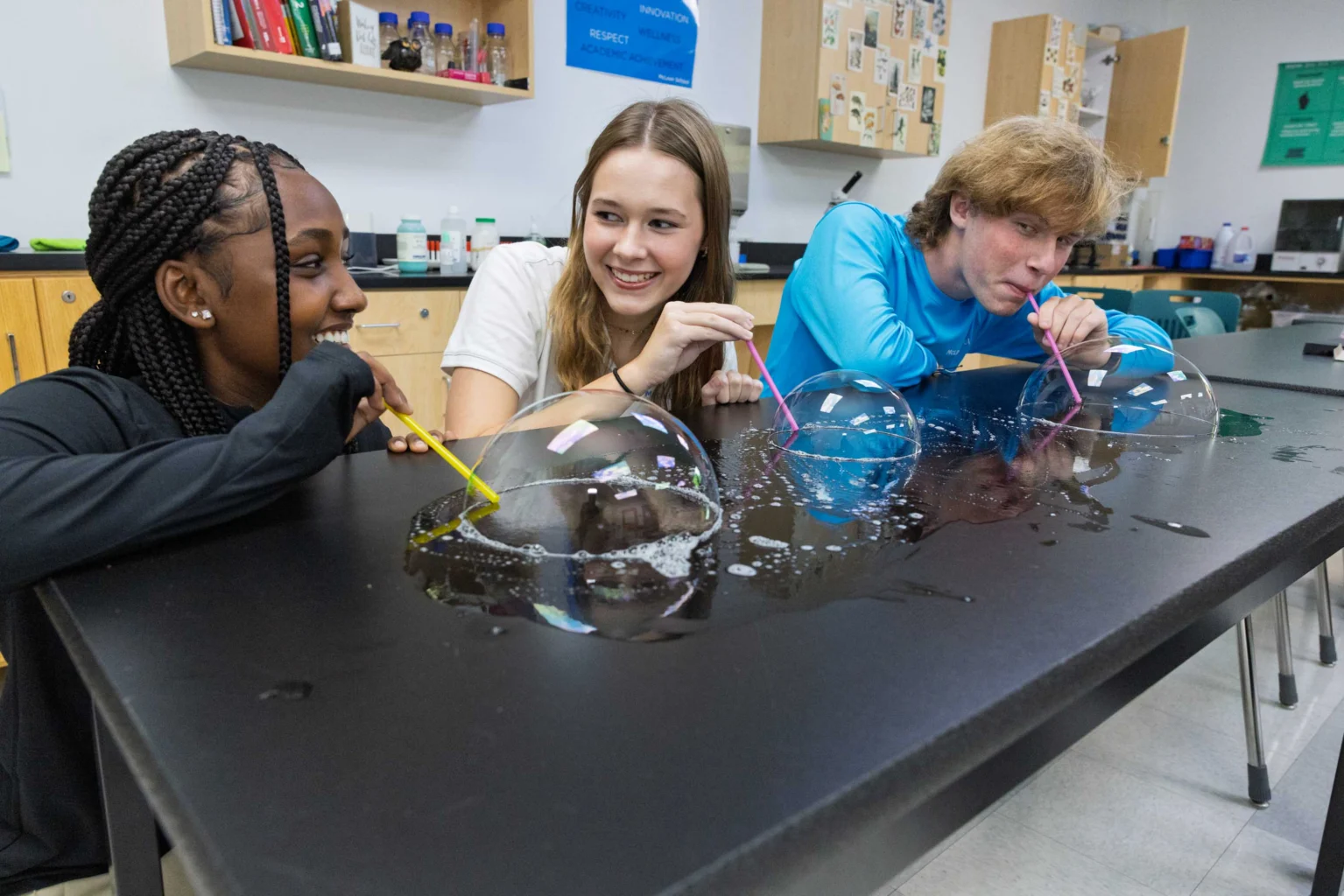 Three students use straws to blow soap bubbles on a black table in a science classroom.
