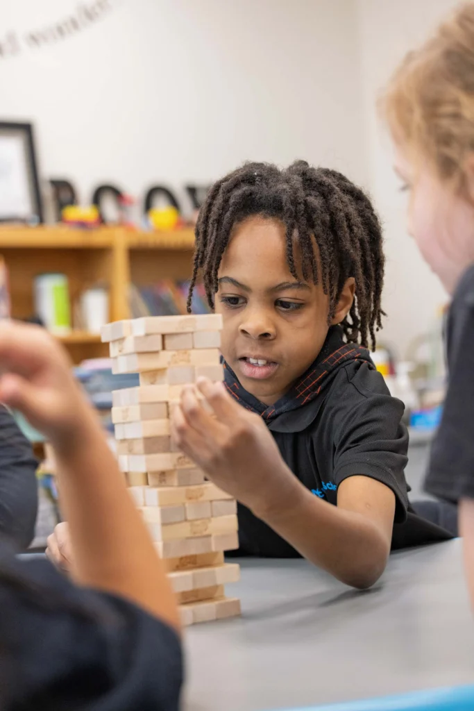 A child focuses on carefully removing a block from a stacked wooden Jenga tower while other children watch.