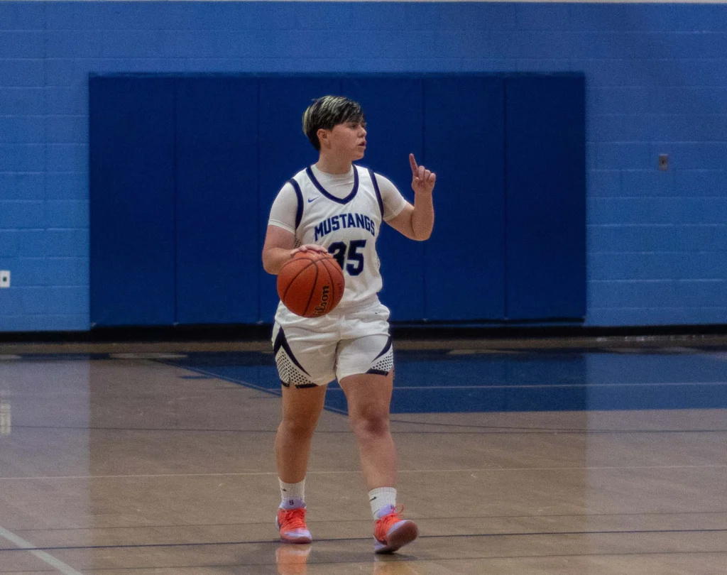 A basketball player in a white "Mustangs" uniform dribbles the ball on an indoor court while raising their left hand and looking to the side.