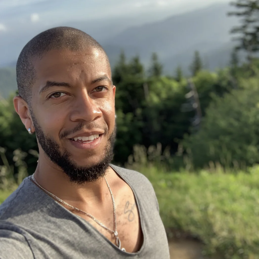 A man with a beard and short hair takes a selfie outdoors with trees and mountains in the background on a sunny day.