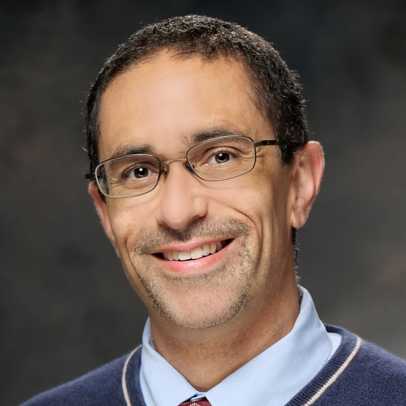 A man with short curly hair, glasses, and a trimmed mustache and beard is smiling, wearing a blue sweater over a collared shirt and tie, with a dark, blurred background.