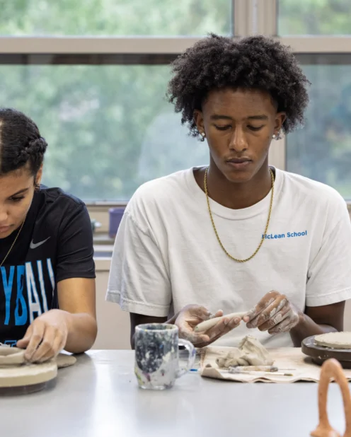 Two students sit at a table working on clay projects, each focused on shaping clay with their hands in a classroom setting.