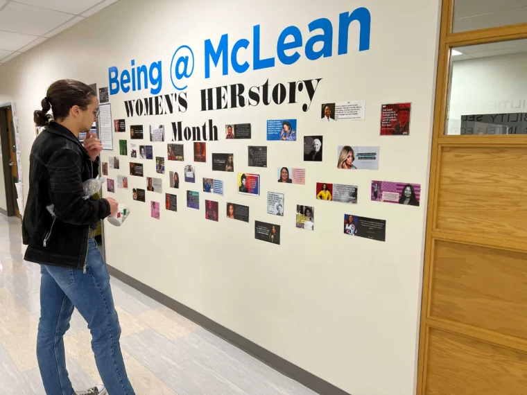 A person stands in a hallway looking at a wall display celebrating Women's HERstory Month with photos and information about notable women under the heading "Being @ McLean.