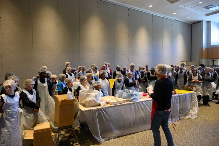 A large group of people wearing hairnets and aprons listen to an instructor in a conference room with tables and packing materials.