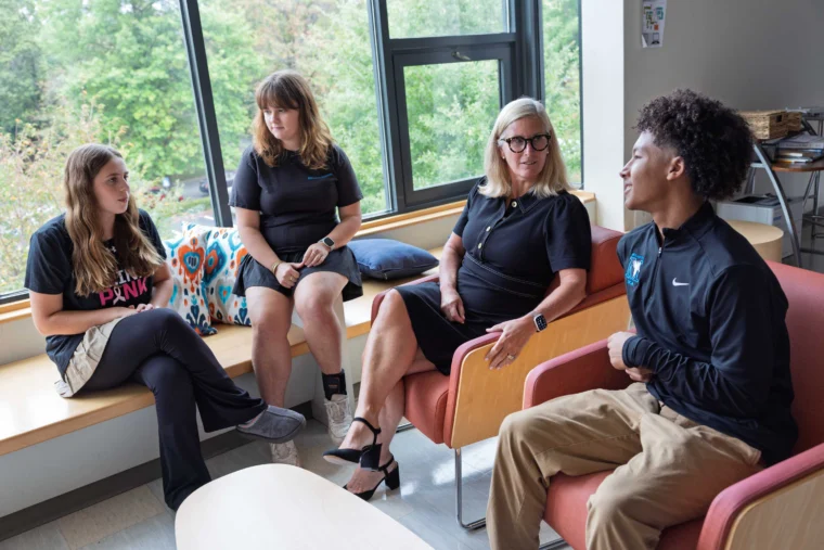 Four people sit and talk in a brightly lit room with large windows and trees visible outside. Three women and one man are seated, engaged in conversation.