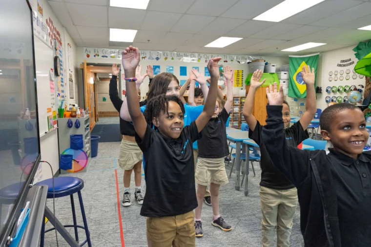 A group of elementary school children stand in a classroom with their arms raised, participating in an activity together.