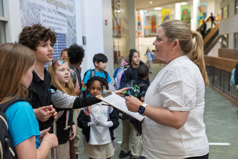 A teacher holding a clipboard speaks to a group of elementary school students gathered in a hallway.