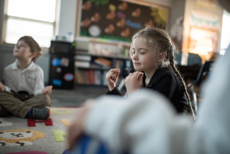 Children sit cross-legged on a classroom rug, eyes closed and hands raised, appearing to practice mindfulness or meditation.