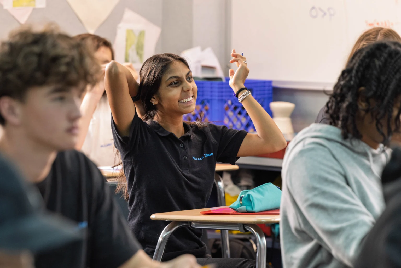 A student smiles and raises her hand in a classroom while other students sit at their desks, listening attentively.