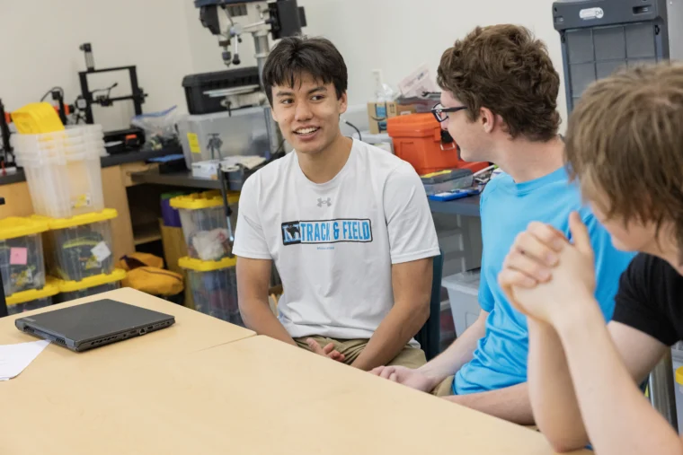 Three young men sit around a table in a workshop or classroom, engaged in conversation. Tools and storage bins are visible in the background.
