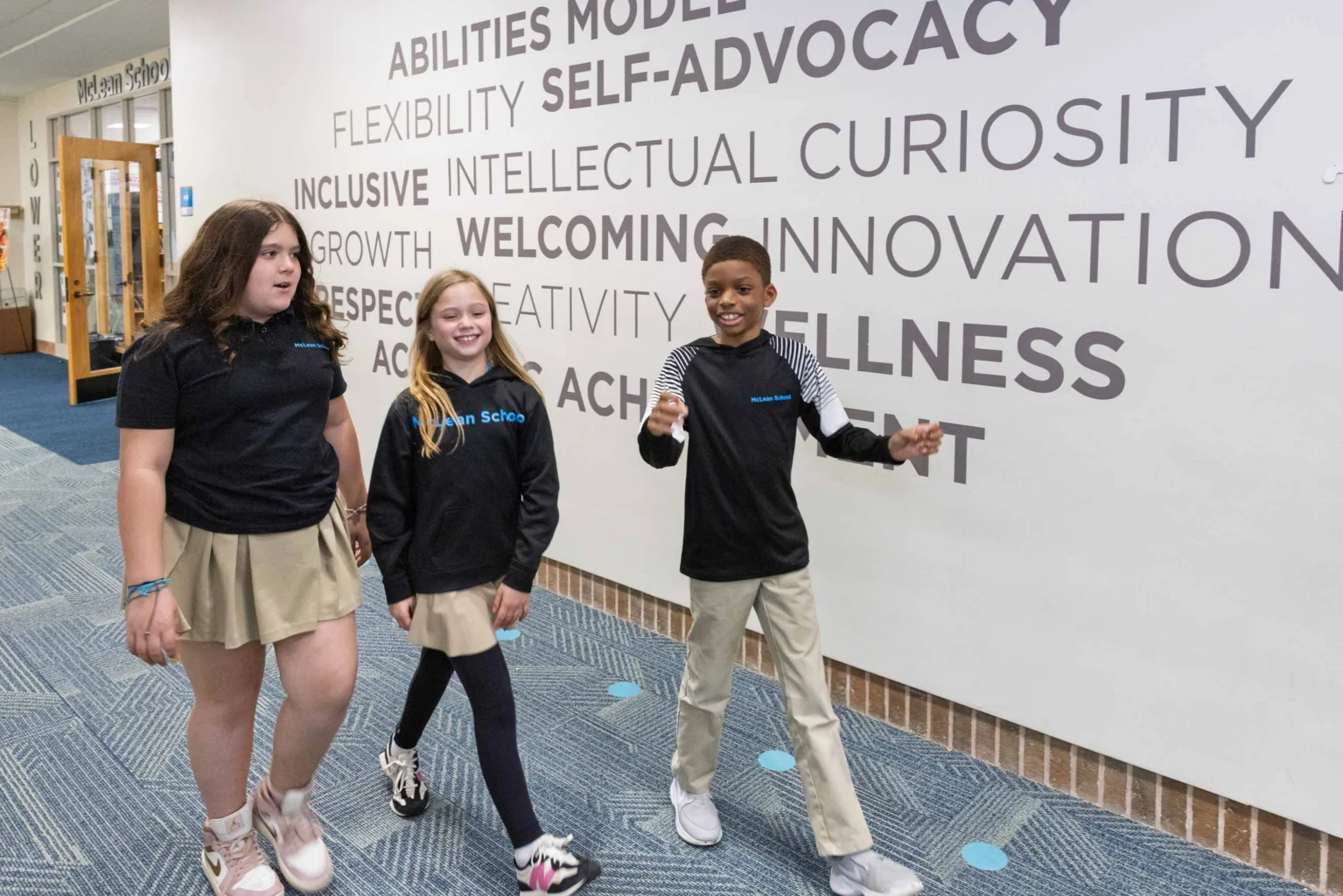 Three students in school uniforms walk and talk in a hallway with motivational words like "self-advocacy," "innovation," and "wellness" displayed on the wall behind them.