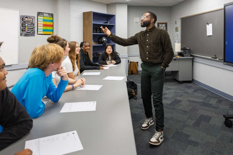 A man stands and gestures while speaking to a group of students seated at a classroom table with papers in front of them.