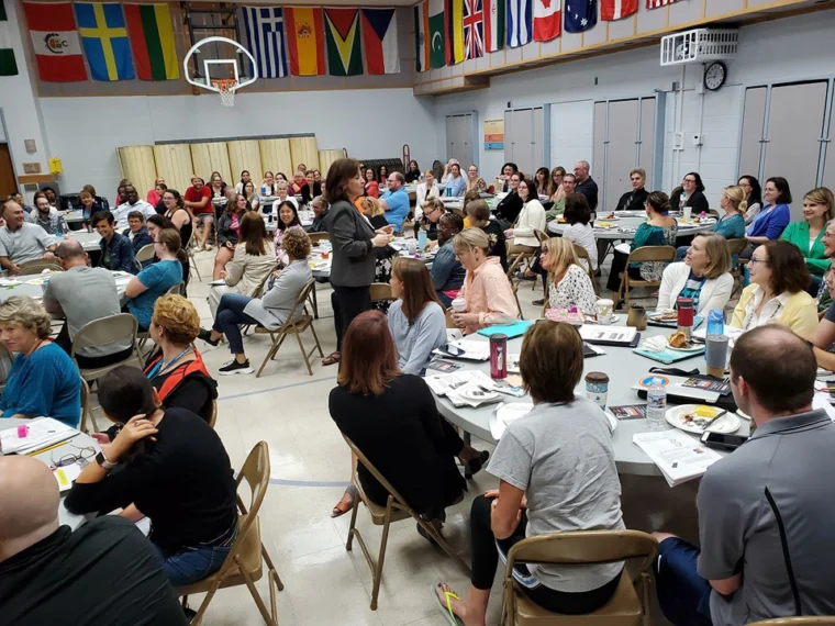 A large group of adults sit at round tables in a gymnasium, listening to a woman speaking at the center; international flags are displayed on the wall.