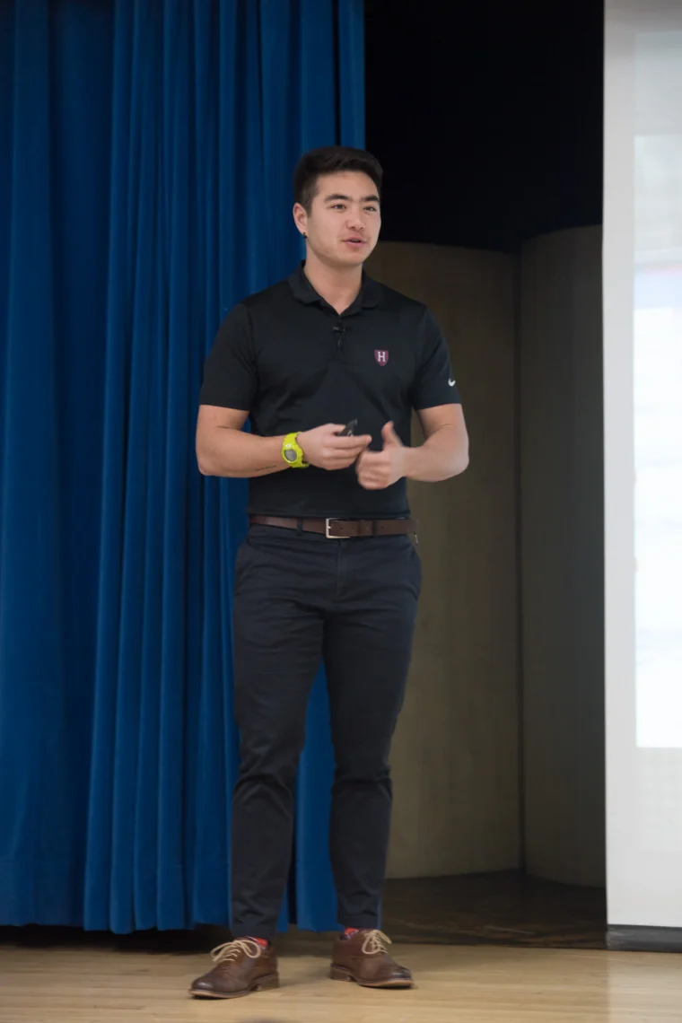 A man in a black shirt and dark pants stands on a wooden floor, speaking in front of a blue curtain and a projection screen.