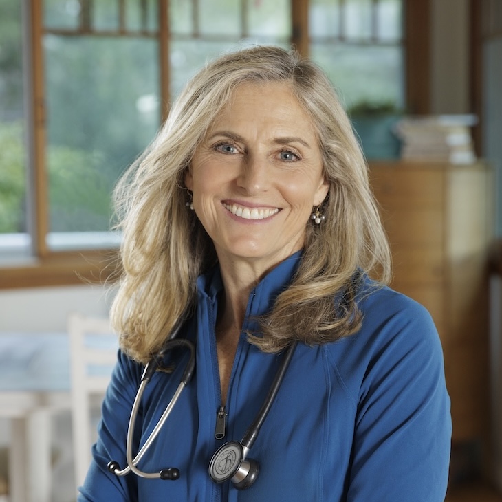 A woman with long blond hair wearing a blue top and a stethoscope smiles at the camera in a well-lit room with a window in the background.