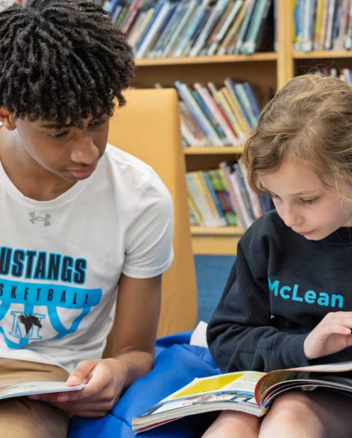 A teenage boy and a young girl sit together in a library, each reading a book. Shelves filled with books are visible in the background.