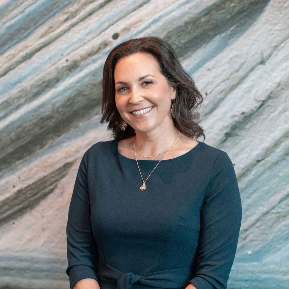 A woman with shoulder-length brown hair in a dark green dress poses and smiles in front of a textured stone wall.