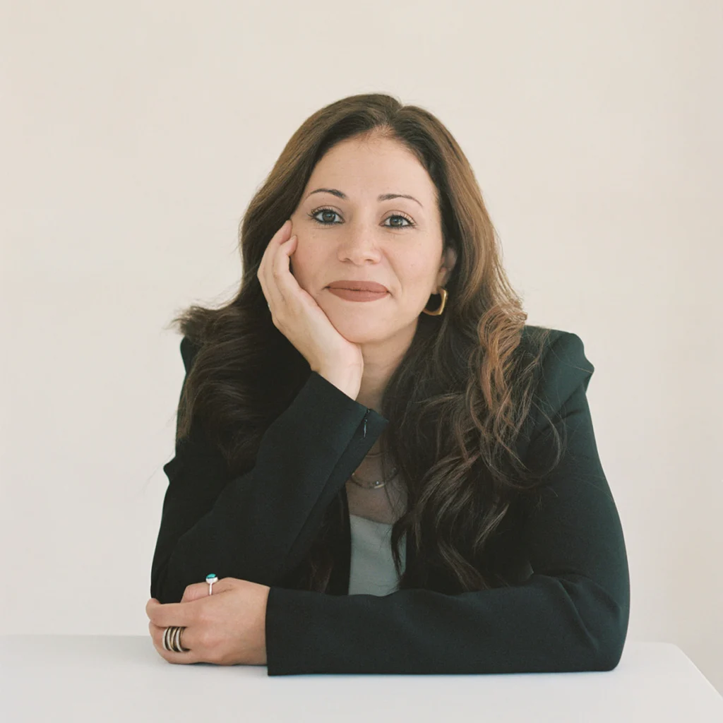 A woman with long brown hair, wearing a black blazer and hoop earrings, sits at a white table, resting her chin on her hand and looking at the camera.