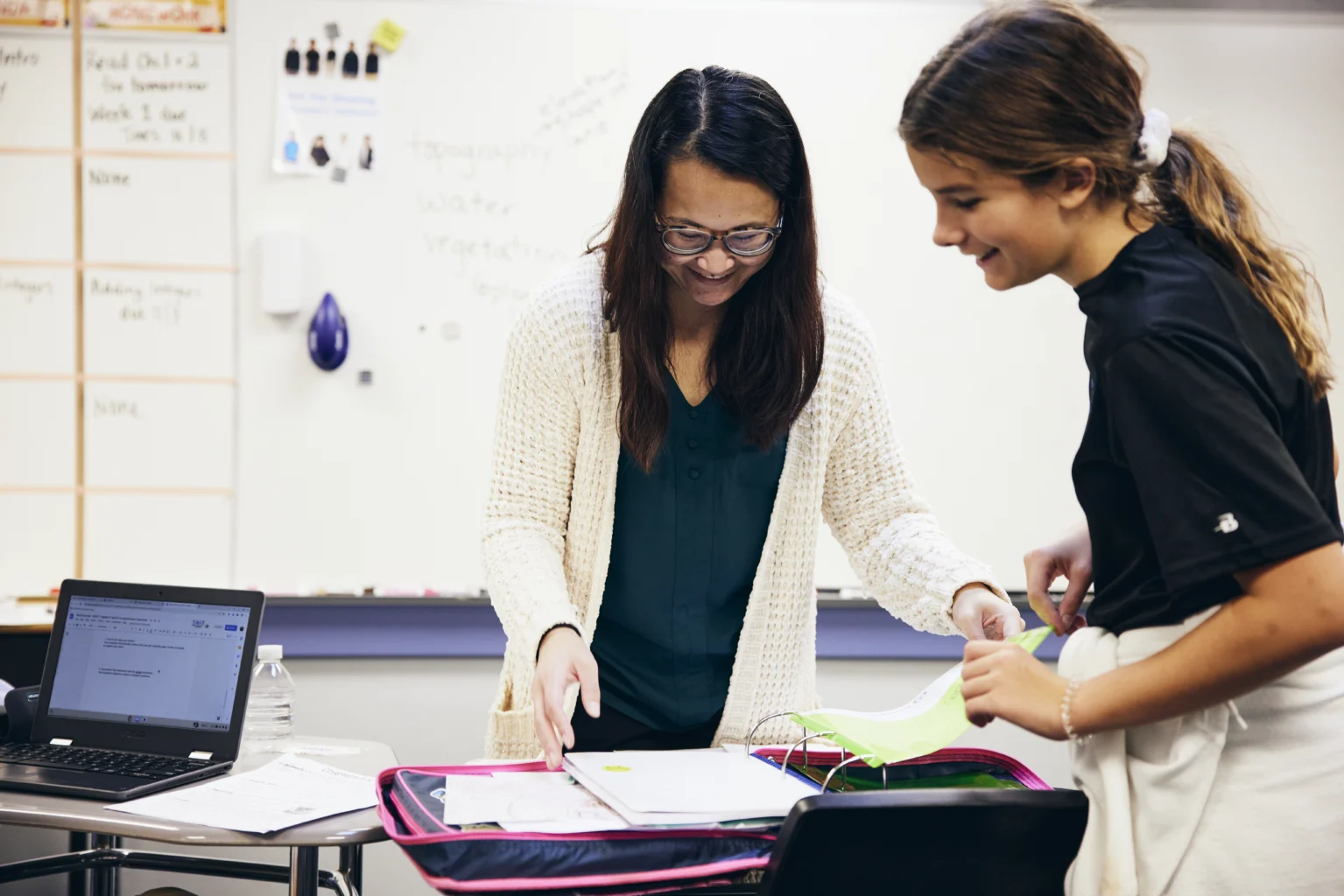 A teacher and a student review papers together at a desk in a classroom with a laptop and a whiteboard in the background.