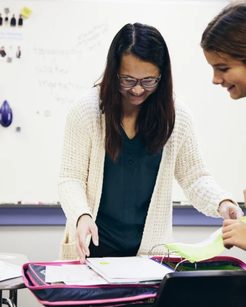 A teacher and a student review papers together at a desk in a classroom with a laptop and a whiteboard in the background.