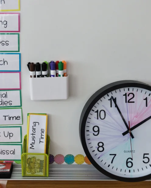 Classroom schedule with time slots and subjects is displayed on a whiteboard next to a round wall clock showing 10:09 and a holder with colored markers.